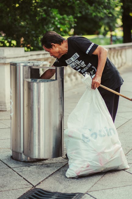 A middle-aged man with dark hair, wearing a black T-shirt with printed text and dark pants, is seen disposing of rubbish into a tall, cylindrical stainless steel public litter bin. The bin features a brushed metal finish with a semicircular opening at the top, designed for waste collection. He is bent forward slightly, holding a large white plastic bag, which appears to be filled with rubbish, in his left hand, while using his right hand to drop waste into the bin's opening. The scene takes place outdoors on a paved surface, possibly a sidewalk or park pathway, bordered by a low stone wall with decorative balustrades on the right side. In the background, lush green trees and foliage are visible, indicating a pleasant environmental setting. The natural lighting and shadowing suggest daytime, and the overall scene depicts an example of individual or community waste disposal, relevant to private rubbish removal services like those provided by Waste Collection Edgware, in an environment where alternative waste handling methods may be employed.