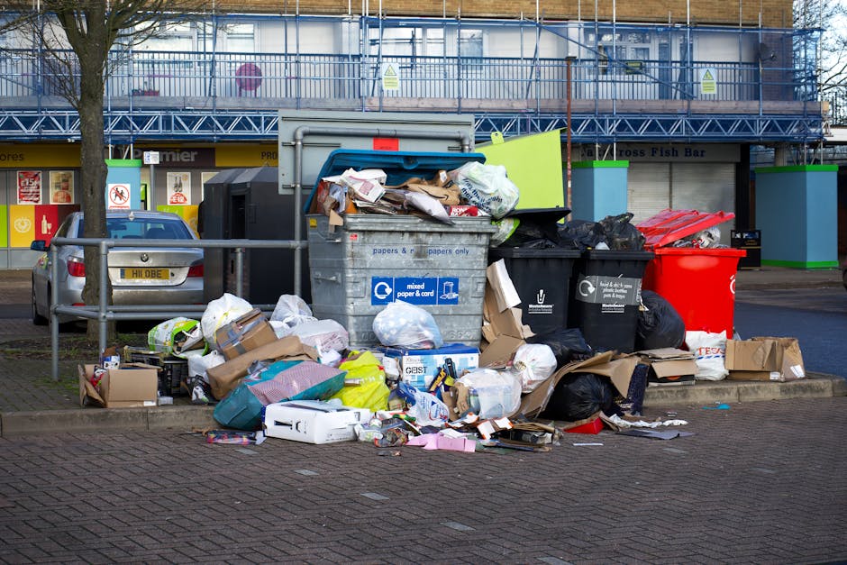 A pile of mixed household rubbish and discarded packaging materials collected on a paved urban street, with cardboard boxes, plastic bags, and loose paper scattered in front of a large metal bin labeled for mixed paper and card. Several black and red rubbish bags are stacked beside the bin, which is partially overflowing with unwrapped waste. Behind the pile, there is a parked grey car positioned close to a metal barrier, with a background of a commercial building featuring storefronts and advertisements. The scene is outdoors under natural daylight with some trees visible, and a construction scaffold is present above the building indicating possible renovation work. The overall composition emphasizes a typical refuse accumulation scenario that might be addressed through private waste collection services like Waste Collection Edgware, aligning with the context of alternative rubbish disposal options outside of municipal collection.