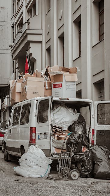 A white cargo van with its rear doors open, parked on a city street in front of a multi-storey building with beige and grey facades. The inside of the van is filled with large black garbage bags, flattened cardboard boxes, and white plastic-wrapped items, indicating a collection of bulky rubbish. Several cardboard boxes, some torn open, are stacked on top of the van, with a few leaning against the vehicle. In front of the open van, on the pavement, there are additional black trash bags and a small hand truck loaded with more waste materials, including a few loose cardboard pieces. The scene suggests an urban environment for independent rubbish removal, with waste typically associated with on-site clearance or private disposal services. Natural lighting highlights the textures of the cardboard, plastic, and vehicle surfaces, emphasizing the cluttered appearance of the rubbish collection, which aligns with alternative waste handling methods outside typical council services.