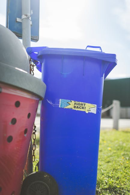 A tall, bright blue plastic waste bin with a closed lid, positioned outdoors on a patch of grass. The bin features a small, rectangular sticker with the words 'FIGHT BACK!' in yellow and black print, partially torn. Next to the blue bin, on its left, is a red waste container with a gray lid, both of which appear to be part of rubbish collection or disposal options. In the background, there is a metal pole, possibly a street sign or lamp post, and a section of fencing or barriers, with a clear sky overhead suggesting daytime. The scene indicates an area intended for waste management, emphasizing an organized approach to rubbish collection, often representing alternative or private waste handling services such as those provided by Waste Collection Edgware. The setting appears clean, with natural lighting casting soft shadows, contributing to an overall neutral and professional presentation of outdoor waste containers used for rubbish removal.