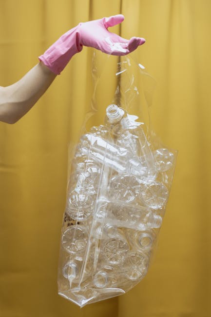 A person wearing a light beige long-sleeve top and pink rubber gloves is holding a large, transparent plastic bag filled with clear, empty plastic bottles and caps. The bag appears to be composed of thin, flexible plastic material and is partially gathered at the top where the person's gloved hand grips it. The background is a solid, muted yellow curtain or wall, providing a neutral setting that emphasizes the recyclable waste. The plastic bottles within the bag vary in size and shape, with some featuring screw-top openings visible at the top. The scene is well-lit, highlighting the glossy surface of the plastic bottles and the transparency of the bag, which relates to waste separation and collection services, such as those offered by Waste Collection Edgware for responsible rubbish disposal and recycling.