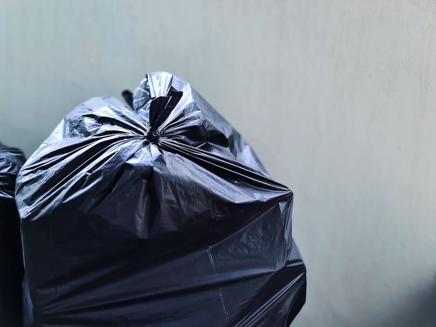 Close-up of multiple black plastic rubbish bags filled with waste, tightly tied at the top, placed against a plain, light-colored wall, likely on a driveway or outdoor area. The bags are glossy and appear to be made of durable plastic, with some slight creases and wrinkles visible on their surfaces. The background shows a neutral wall with minimal texture, providing a simple backdrop that emphasizes the stacked rubbish bags. The scene suggests a private collection or on-site clearance, possibly managed by Waste Collection Edgware, aligning with alternative waste handling methods such as domestic rubbish removal. The lighting is even and natural, highlighting the shine on the plastic surfaces and the contours of the filled bags, creating a clear and detailed visual representation of household waste awaiting disposal or collection.