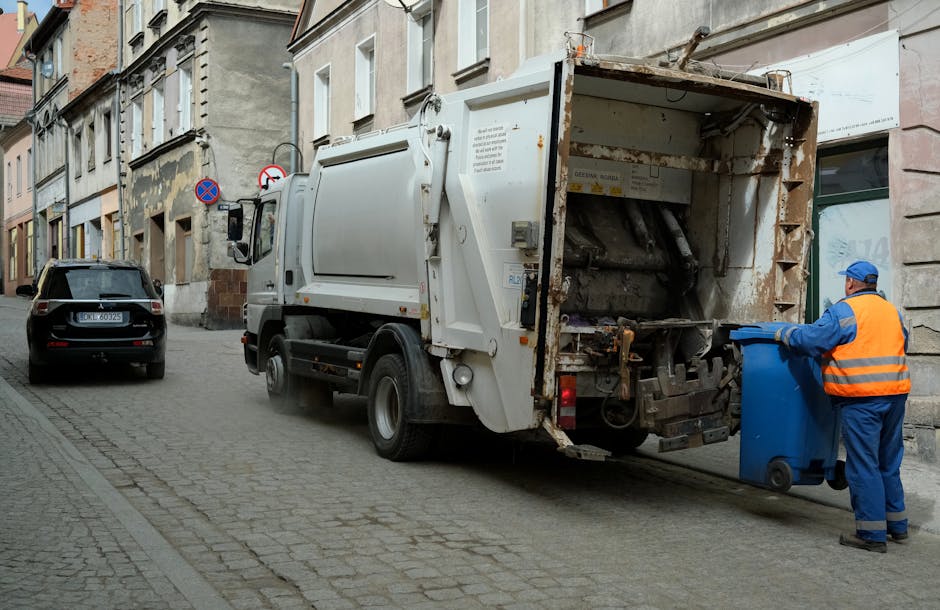 A waste collection truck, white with slight rust and dirt marks, is parked on a narrow urban street beside an old stone and brick building. The truck’s rear compartment is open, revealing mechanical parts and a conveyor system used for collecting rubbish. A worker dressed in a blue uniform and high-visibility orange vest is positioned on the right, emptying a blue wheeled bin into the truck’s bin opening. The street surface is cobblestone, with a black car parked ahead of the truck on the left side. In the background, there are a few street signs mounted on the building wall, including no parking and no stopping signs. The surrounding environment features weathered building facades with worn paint and exposed brickwork, indicating an older city area. Overcast natural lighting creates soft shadows, highlighting the practical nature of the waste removal activity, which is part of a private rubbish collection service, such as Waste Collection Edgware, operating in urban residential zones.