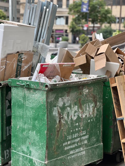 A large, weathered green waste skip positioned on a paved outdoor area, filled with various types of debris including cardboard boxes, some flattened and others intact, along with crumpled paper and plastic wrapping. To the left of the skip, there are several metal rods or frames bundled together, leaning against it, while adjacent to the right, there are additional cardboard and wooden materials, some partially crushed or deconstructed. In the background, blurred cityscape elements such as buildings, trees, and parked vehicles are visible under natural daylight, suggesting an urban setting suitable for private waste disposal or alternative rubbish collection services, as managed by companies like Waste Collection Edgware. The overall impression is of a typical scene representing on-site clearance or bulk rubbish removal, with the focus on the collection of mixed waste materials from residential or commercial premises in London boroughs.