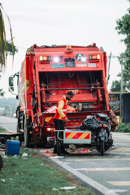 A waste collection truck, white with slight rust and dirt marks, is parked on a narrow urban street beside an old stone and brick building. The truck’s rear compartment is open, revealing mechanical parts and a conveyor system used for collecting rubbish. A worker dressed in a blue uniform and high-visibility orange vest is positioned on the right, emptying a blue wheeled bin into the truck’s bin opening. The street surface is cobblestone, with a black car parked ahead of the truck on the left side. In the background, there are a few street signs mounted on the building wall, including no parking and no stopping signs. The surrounding environment features weathered building facades with worn paint and exposed brickwork, indicating an older city area. Overcast natural lighting creates soft shadows, highlighting the practical nature of the waste removal activity, which is part of a private rubbish collection service, such as Waste Collection Edgware, operating in urban residential zones.