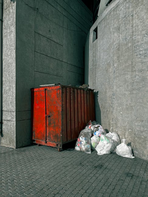 A red metal skip with a weathered, slightly rusted surface is positioned against a dark green wall and a light grey concrete wall at the corner of an outdoor urban area. The skip is rectangular with vertical ribbed sides and a flat top, showing signs of wear and exposure to the elements. Next to the skip, there is a small pile of white and semi-transparent plastic rubbish bags filled with mixed waste, some lying on their sides and partially open, revealing crumpled contents. The surface beneath the skip and rubbish bags is paved with dark grey interlocking bricks, providing a solid ground for waste collection. In the background, there is a narrow passageway between the walls, with a small, square opening or window in the green wall, and minimal natural light depicted, contributing to an overall utilitarian and somewhat neglected appearance characteristic of private waste handling areas. Waste Collection Edgware’s presence for independent rubbish collection is implied by the scene's setup and waste disposal arrangement.