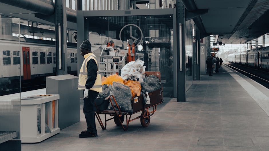 A sanitation worker wearing a black beanie, glasses, a high-visibility vest, and dark clothing stands next to a large, red metal wheelbarrow filled with various rubbish bags and debris, including black, grey, and orange plastic bags. The wheelbarrow is positioned on a spacious, clean concrete platform at a modern train station with glass walls, metal structures, and a corridor extending to the right. Behind the worker, multiple orange hydraulic lifting machines are mounted against the glass wall, and train carriages with closed doors are visible on the left side of the platform. The environment is well-lit with natural light, and there are a few distant passengers walking along the platform. The scene reflects aspects of private waste collection or on-site clearance services often required for efficient rubbish removal in urban transportation hubs, aligning with independent waste handling practices supported by companies like Waste Collection Edgware.
